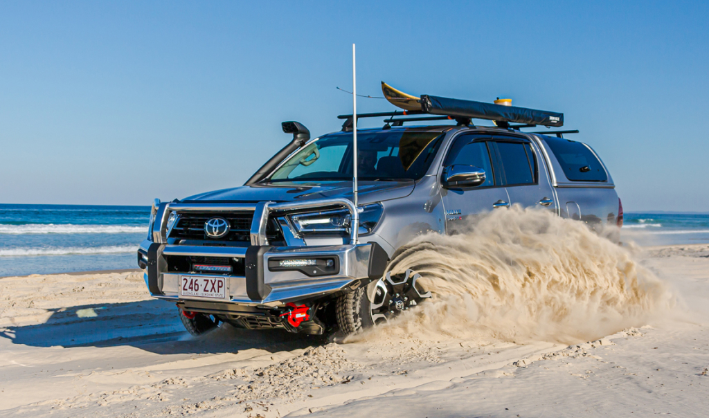 A Toyota HiLux in Ash Slate kicking up a swirl of sand as it drives on the beach. It’s fitted with a Toyota Genuine Winch, Bull Bar, and Roof Rack.