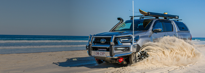 A Toyota HiLux in Ash Slate kicking up a swirl of sand as it drives on the beach. It’s fitted with a Toyota Genuine Winch, Bull Bar, and Roof Rack.