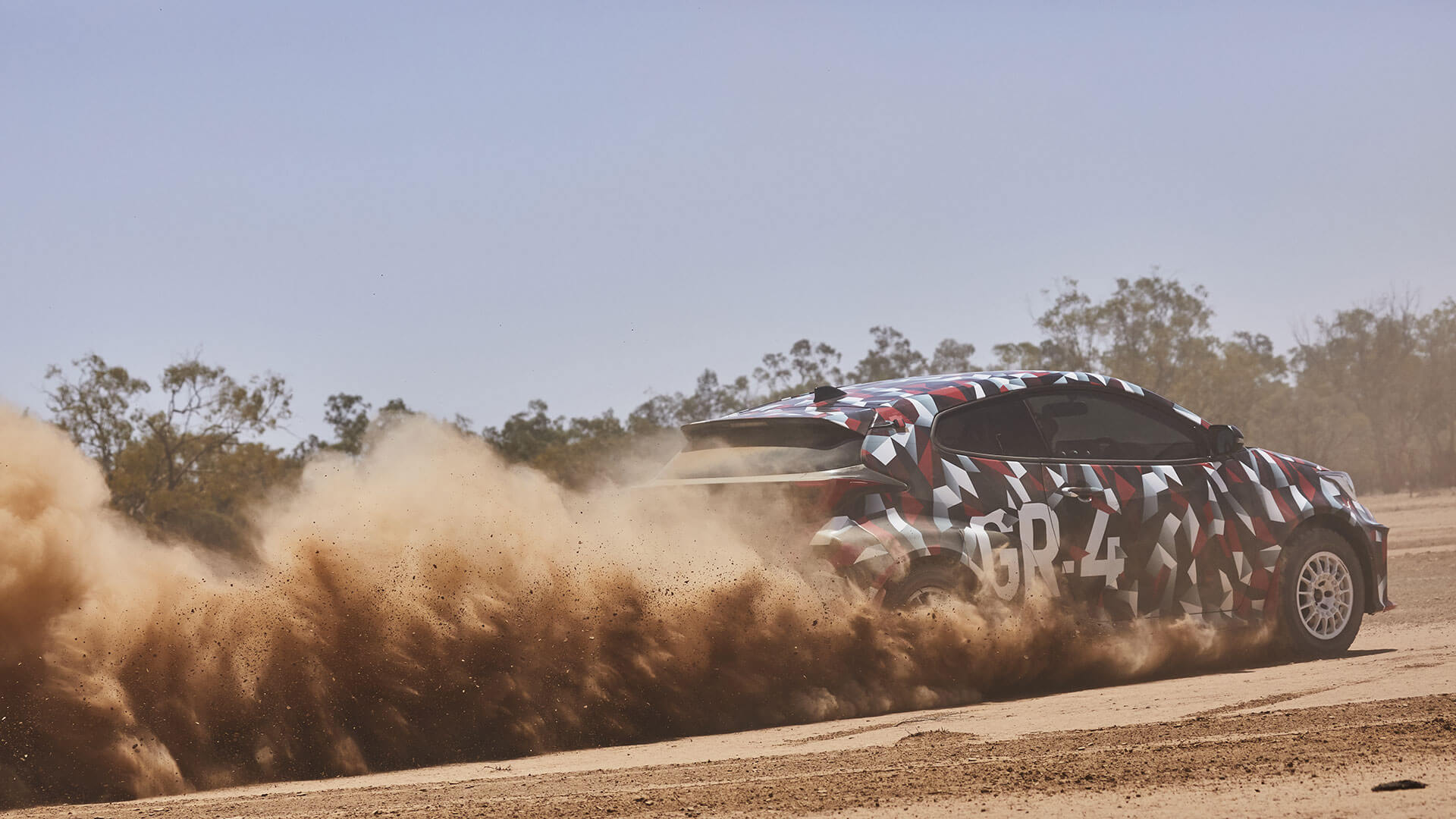 Prototype Toyota GR Yaris shown in camouflage paint driving through a dirt road, kicking up a cloud of dust behind it.