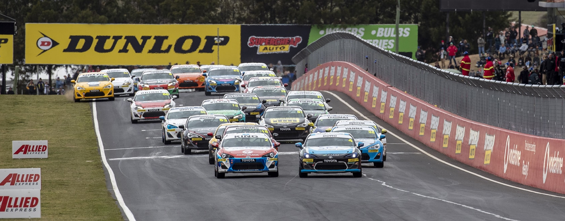 Toyota G R 86s in racing livery compete on a race track lined with spectators watching from behind steel barriers.