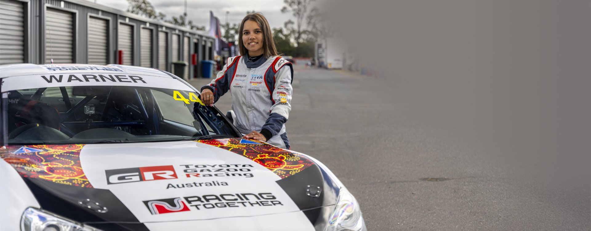 Racing Together's Karlai Warner stands proudly smiling, dressed in protective racing gear, next to a TOYOTA GAZOO Racing-branded GR86.