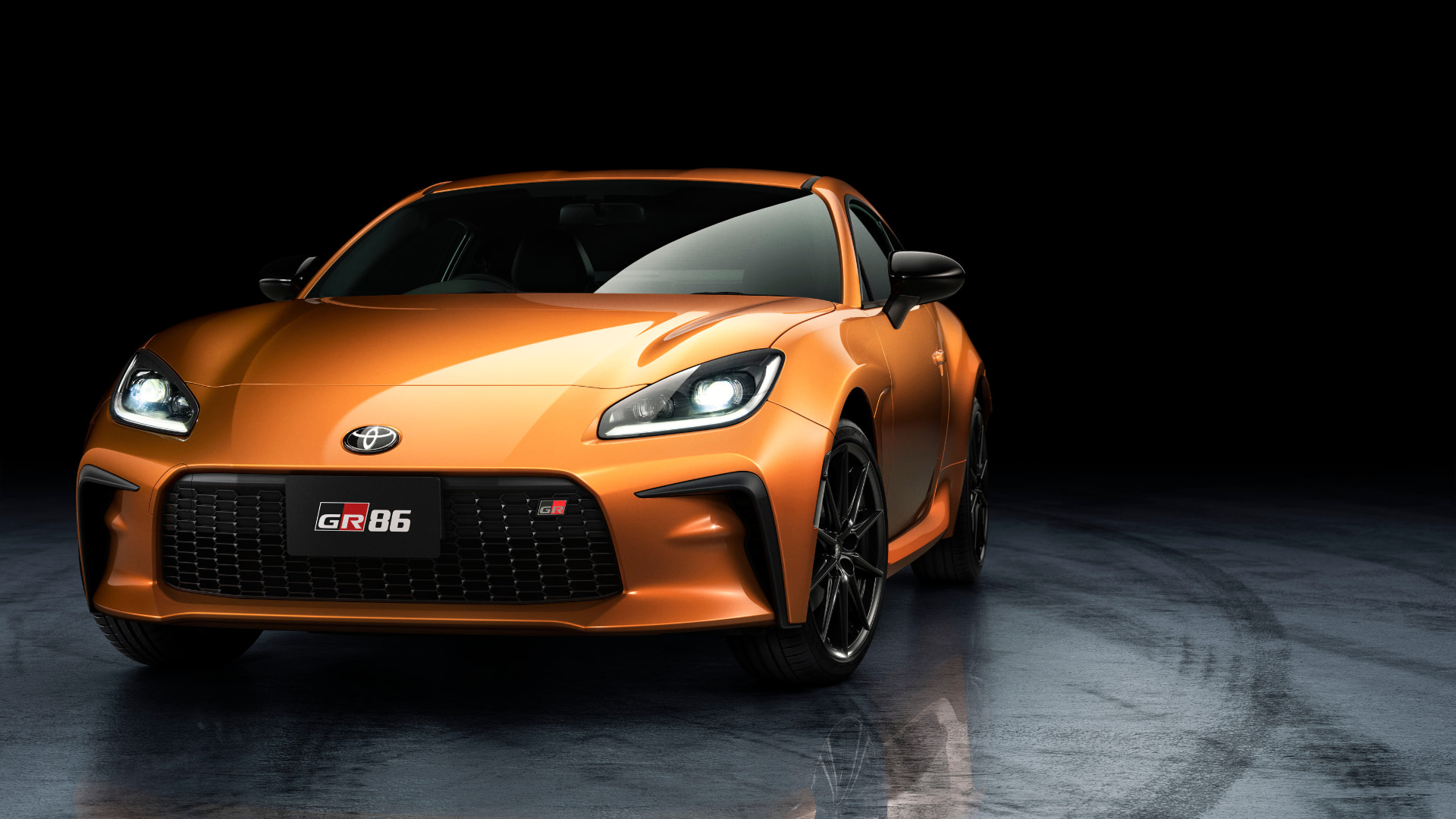 The Toyota GR86 10th Anniversary Edition in Solar Orange, shown in a front three-quarter view in a studio setting with the headlights on, GR86 badging on the grill and black alloy wheels.