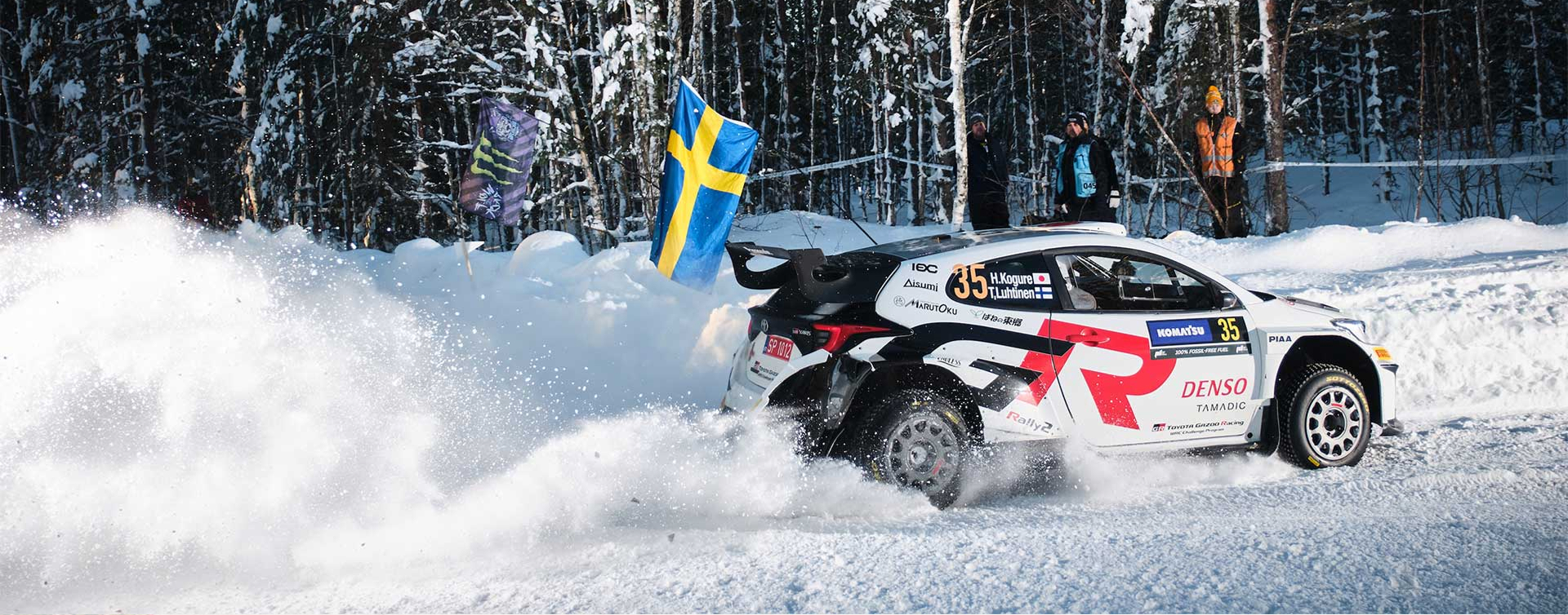 A Toyota G R Yaris Rally2 covered in racing livery decals and flying a Swedish flag. It’s kicking up snow as it drives around a track. Observers are standing in the distance near a line of trees dressed in snow clothing and hi-vis vests.