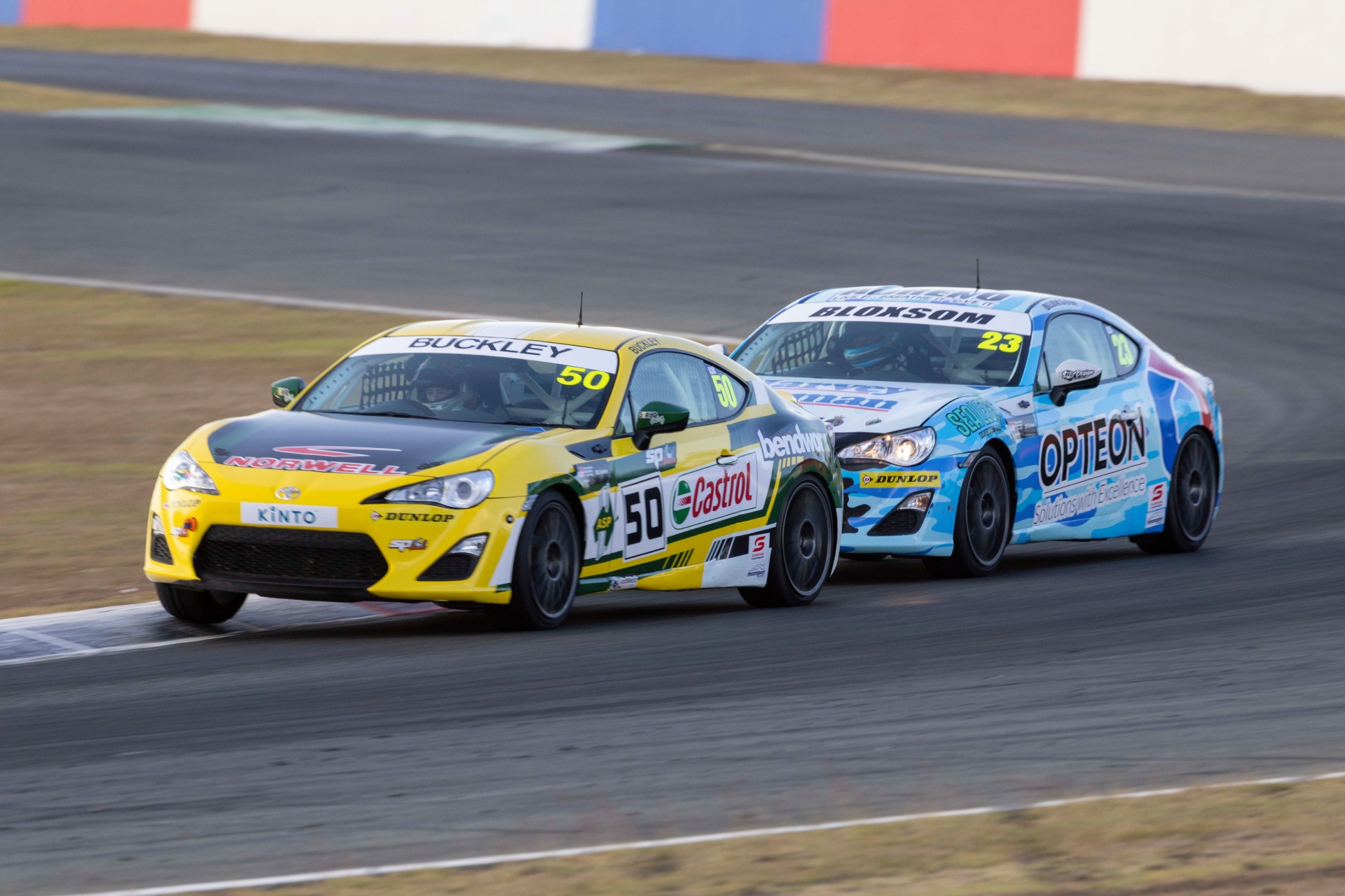 Two Toyota GR 86 race cars, one yellow and one blue, drive close together on a speedway track during the TGRA Scholarship Series.