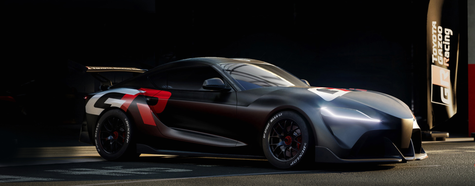 A computer-generated image of a matte grey Toyota G R Supra, with the TOYOTA GAZOO Racing 'G R' logo superimposed on the bonnet and rear wheelhouse. Its L E D headlamp glows against the dark backdrop of a warehouse.