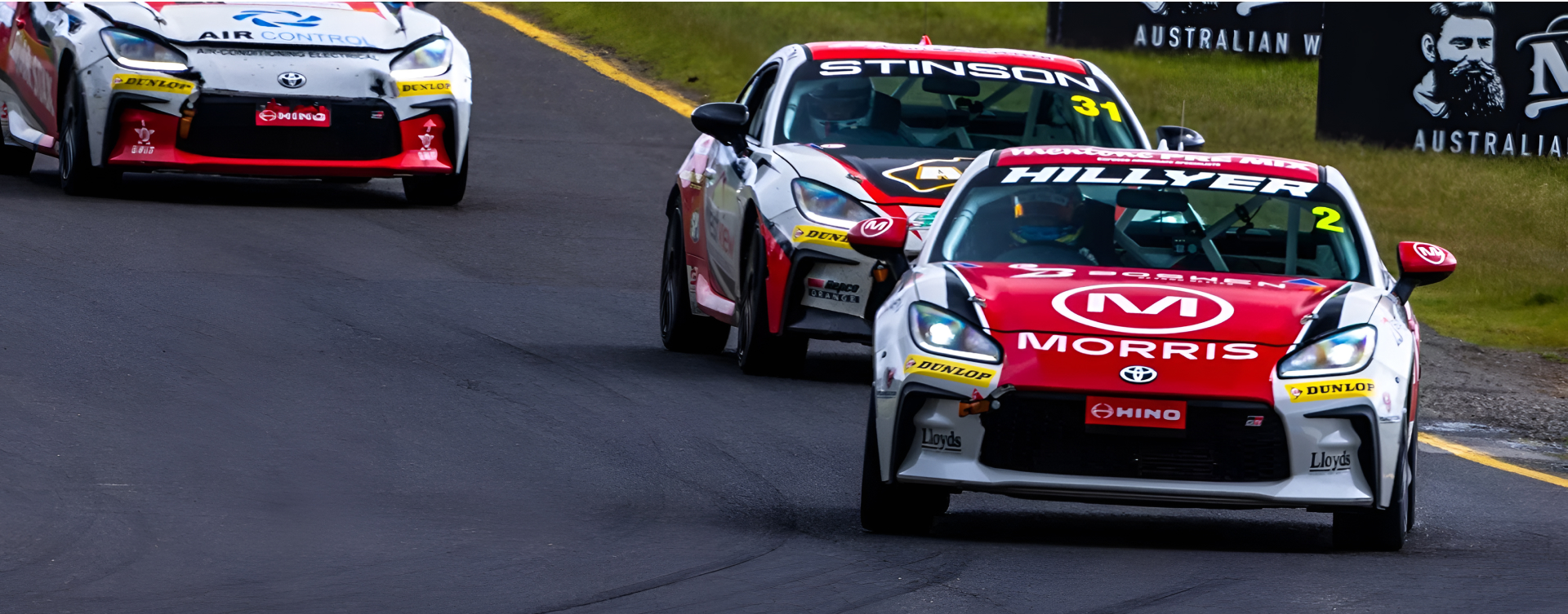 Three GR 86 race cars in red and white racing livery compete at the Sandown 500, the track bordered by grass and event signage.
