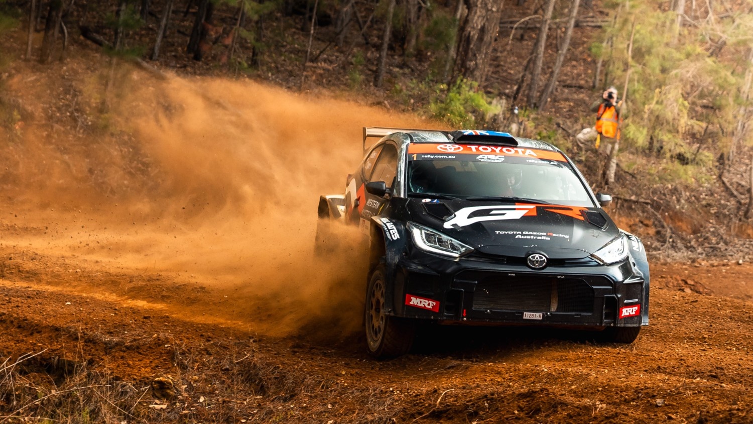 A black Toyota GR Yaris rally car races down a dirt track, kicking up a large cloud of reddish dust.