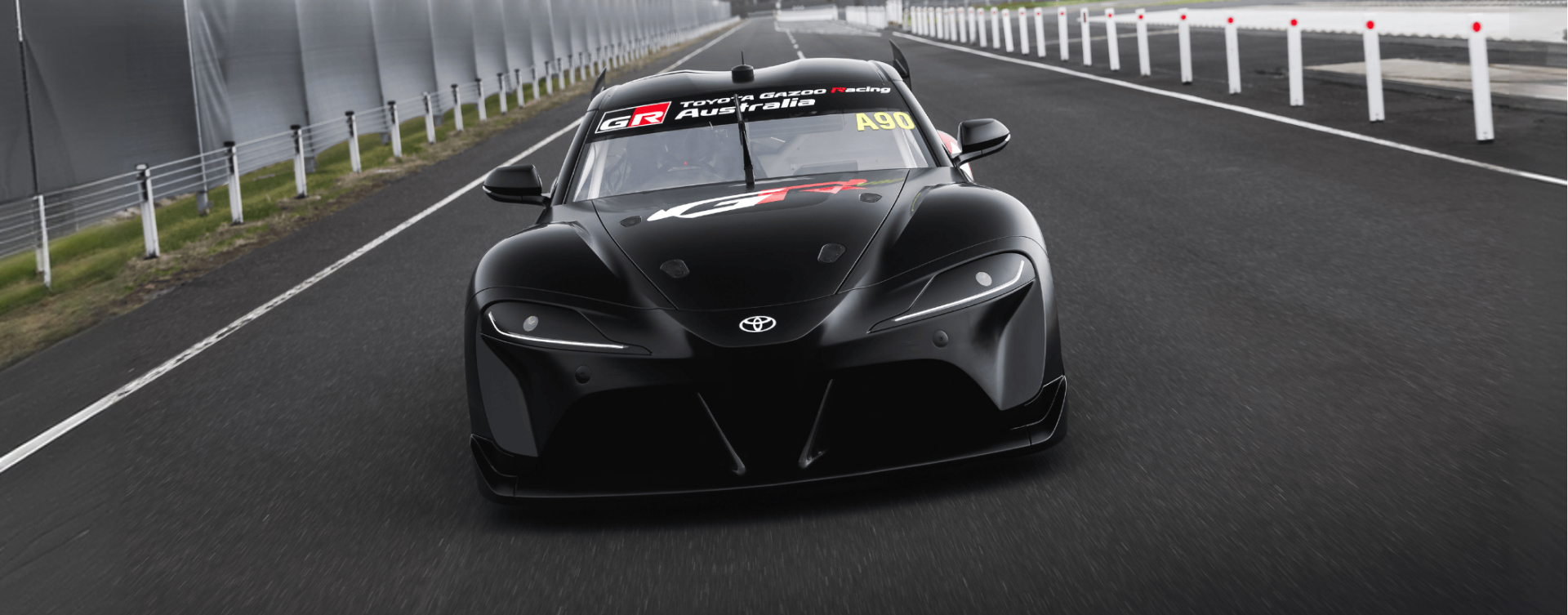 A Toyota GEN3 G R Supra in matte black driving down a clean black racetrack on a foggy day. A TOYOTA GAZOO Racing Australia decal sits above the windshield, with a 'G R' in white and red on the bonnet.