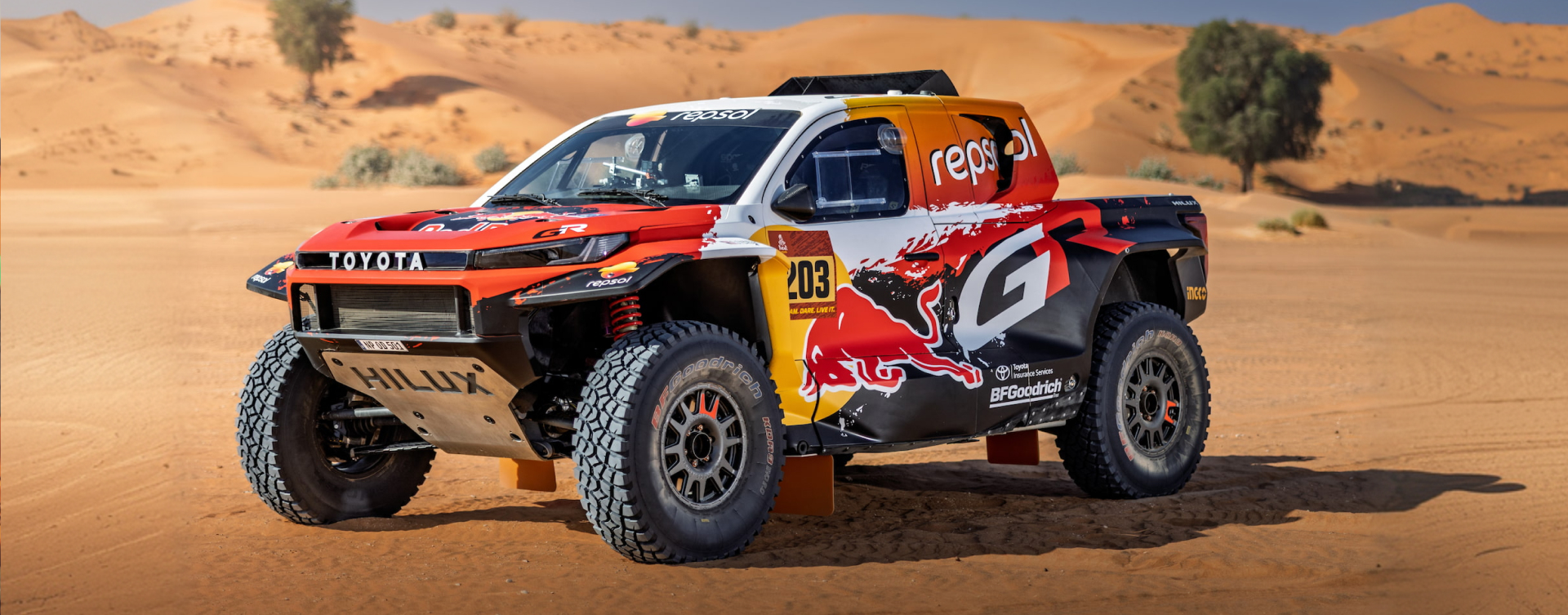 A Toyota Dakar HiLux in the desert, with red, orange and black sponsored livery featuring a G R brandmark. Sparse foliage is scattered around the dunes.   