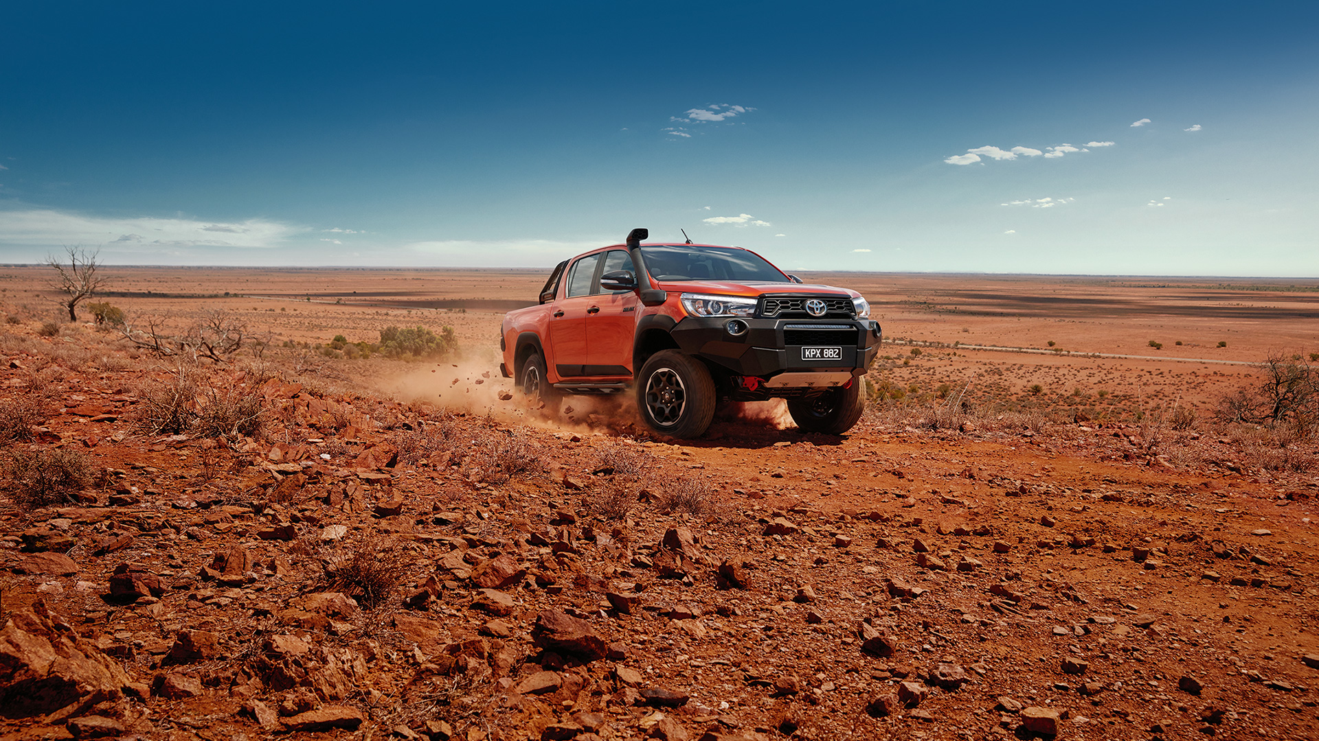 An orange 2018 Toyota Hilux Rogue kicks up the red dirt of Australian outback road, under a beating sun and a clear blue sky.