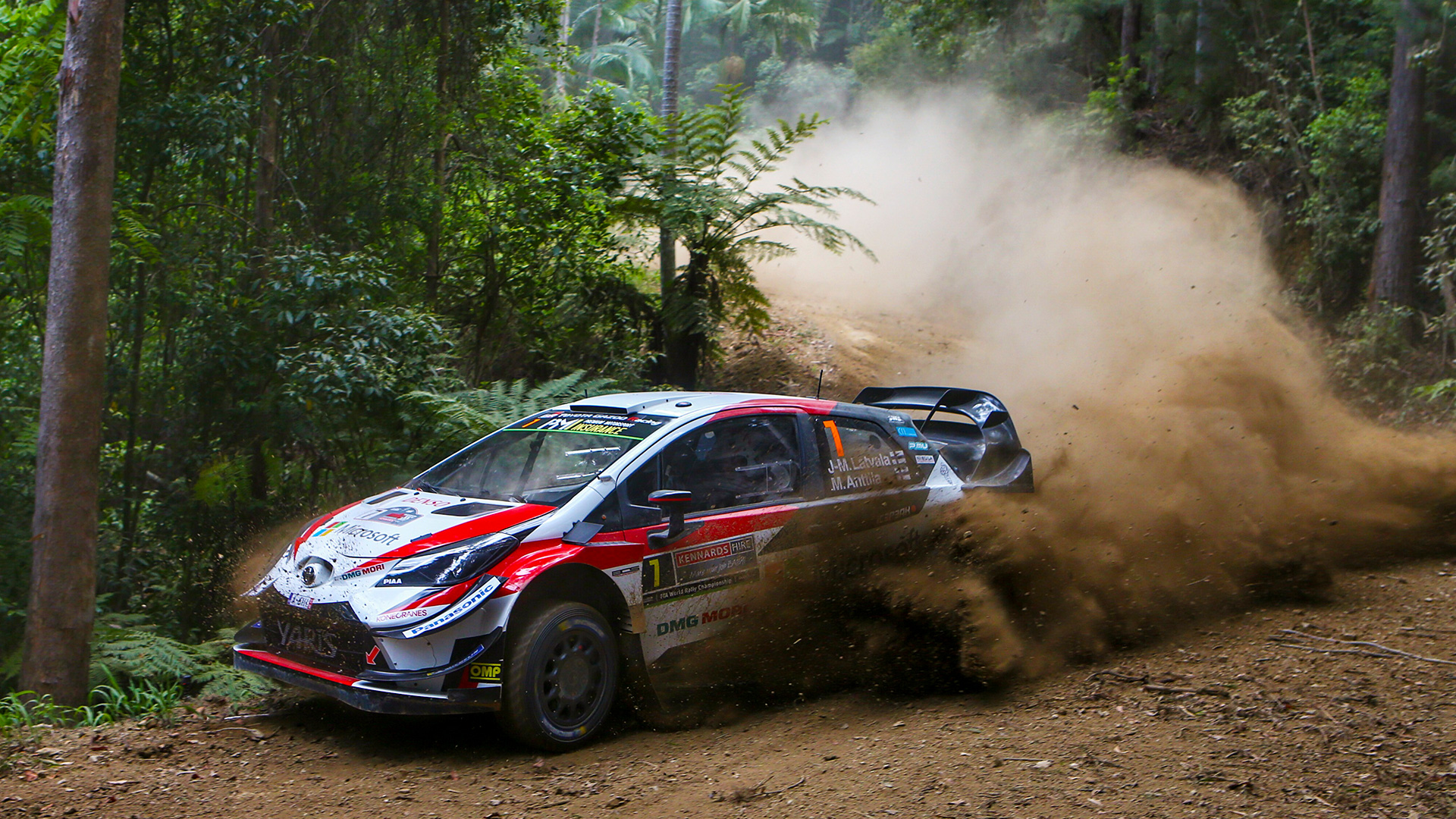 A Toyota Yaris WRC (World Rally Car) drifting around a heavily forested bend, kicking up dust and dirt on the track during the 2018 World Rally Championship.