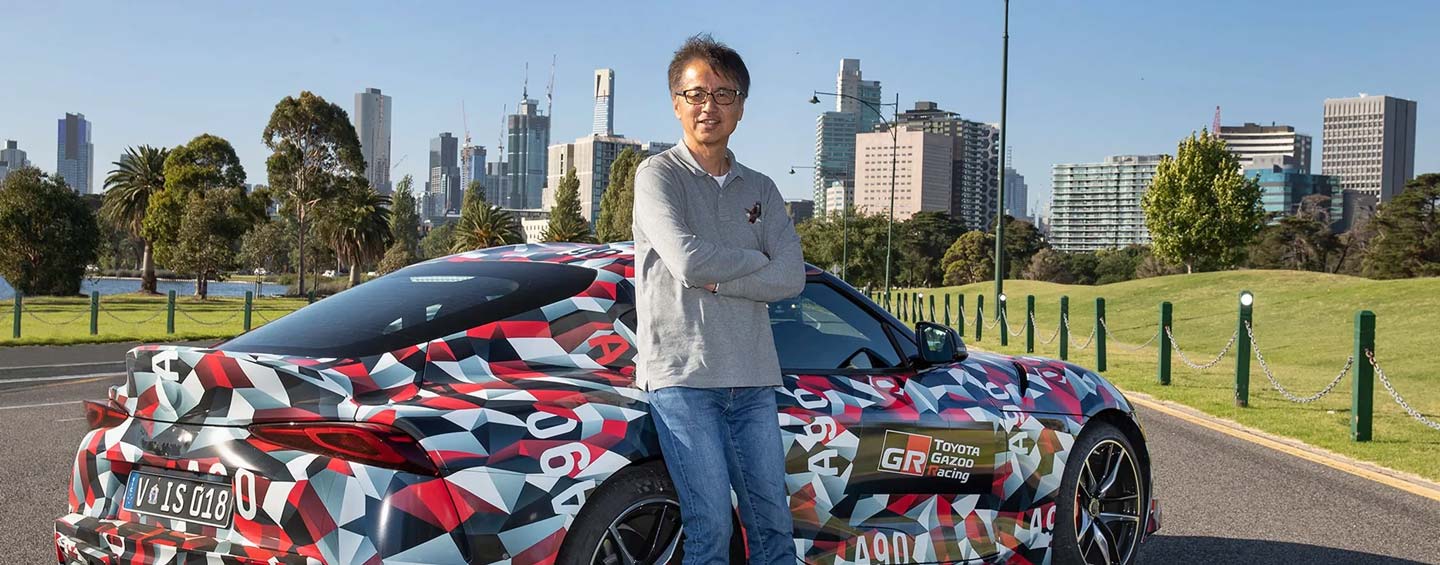 Tetsuya Tada leans against the side of a Toyota GR Supra, with the skyline of Melbourne City in the background.