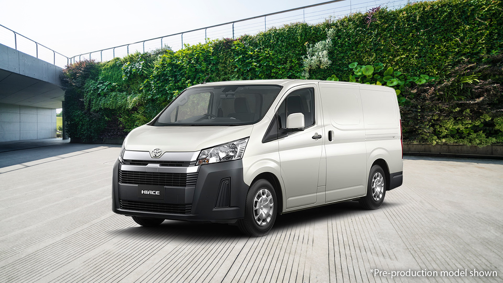 A white Toyota HiAce delivery van is parked on a concrete plaza with a green, leafy wall backdrop.