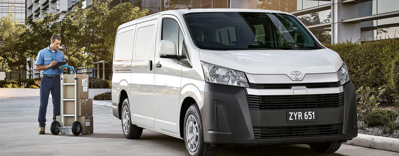 A worker stands examining a digital tablet near a dolly with boxes loaded onto it, near a white 2019 Toyota HiAce.