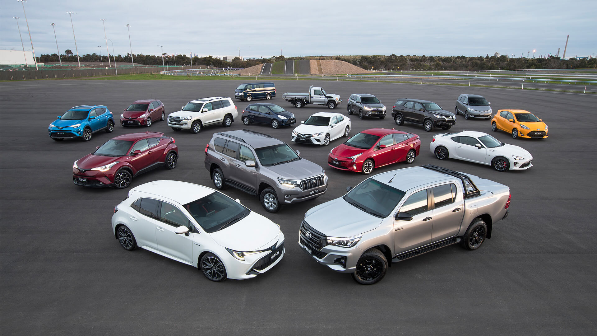 17 different models of Toyota of different colours, all parked together on a large open tarmac.