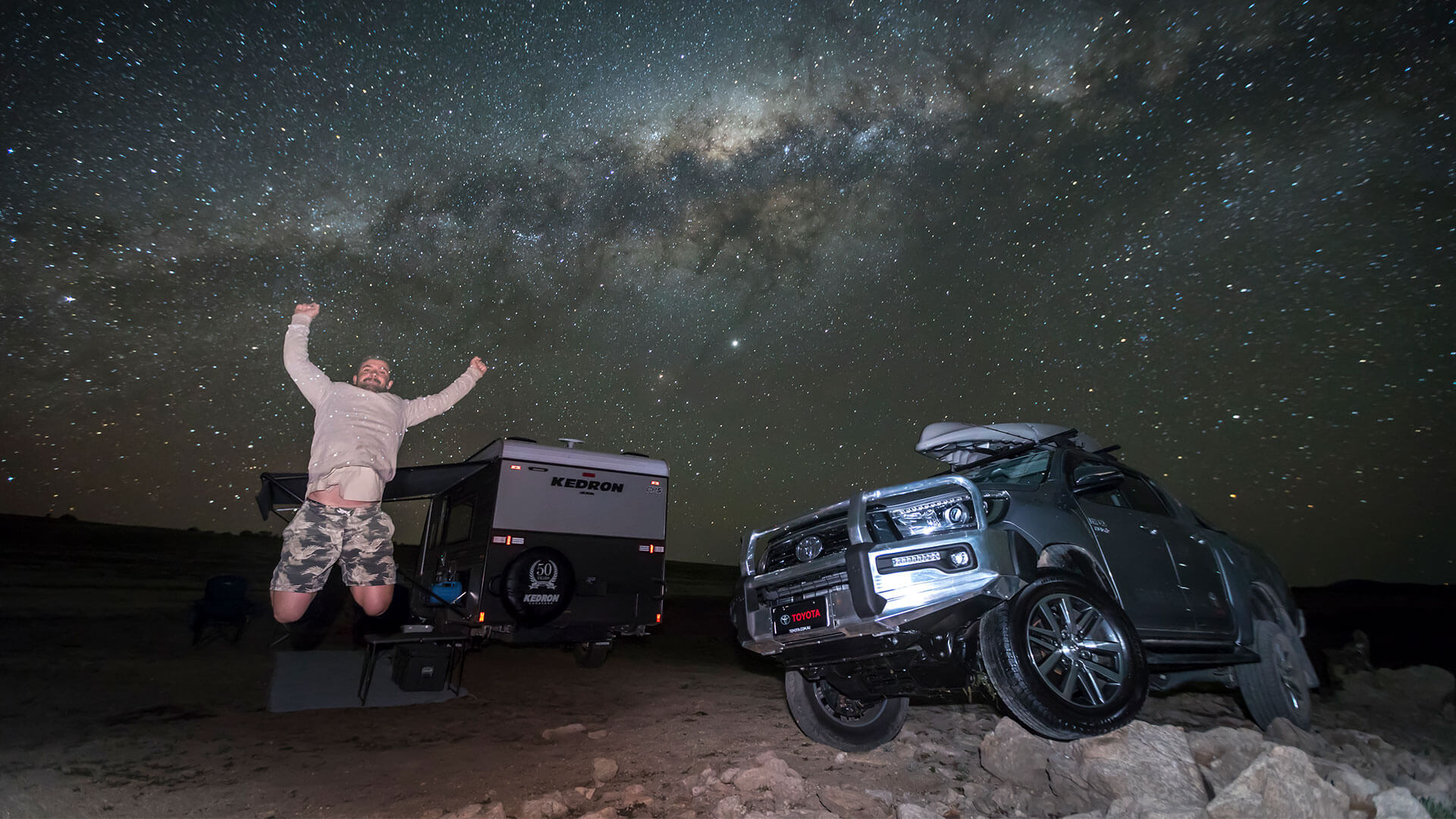 A man jumping for joy in front of a camper trailer and a Toyota Hilux at night, under a sky full of stars.