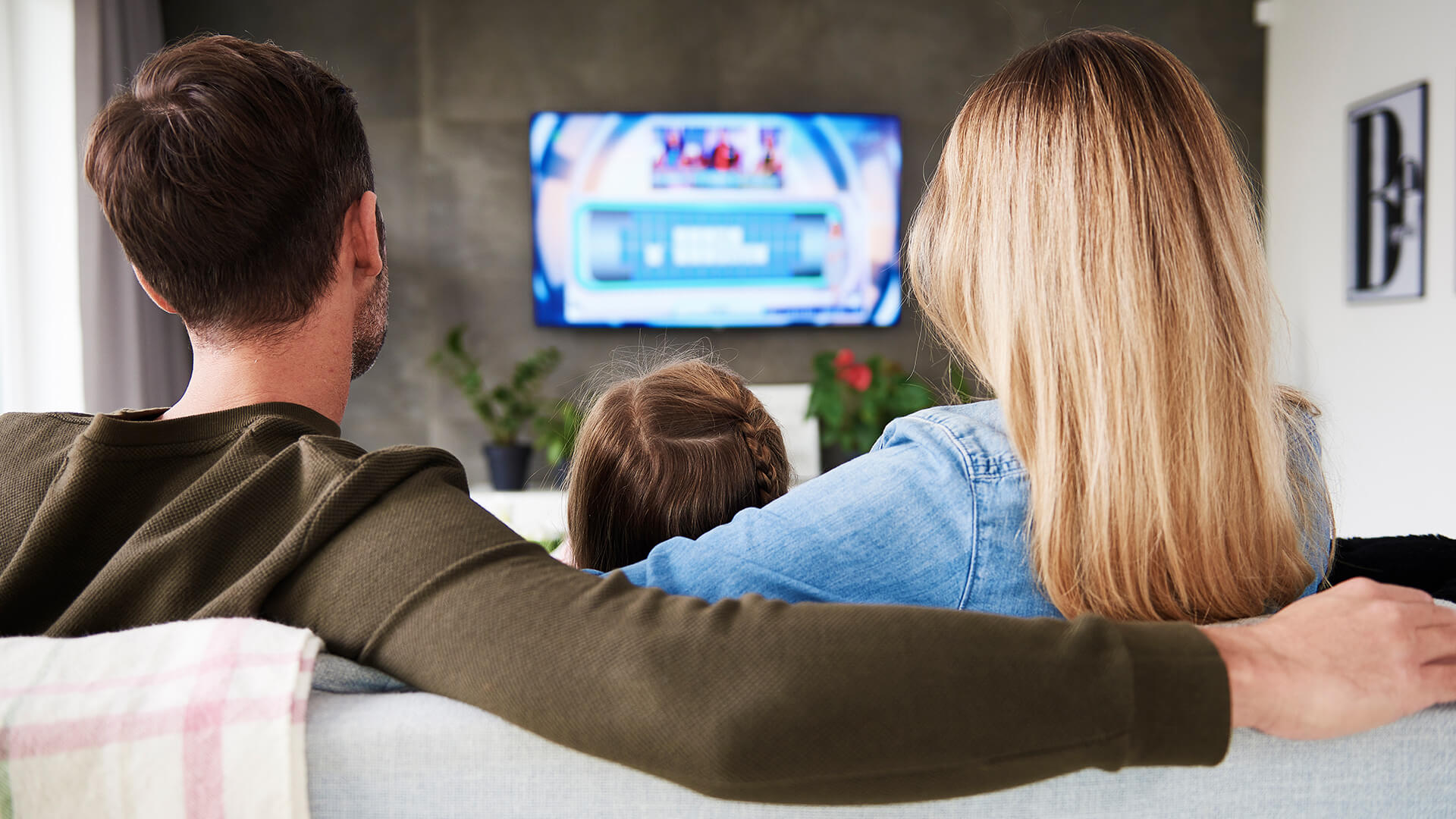 A family of three sit on a couch together in their lounge room, arms around one another, watching their television.