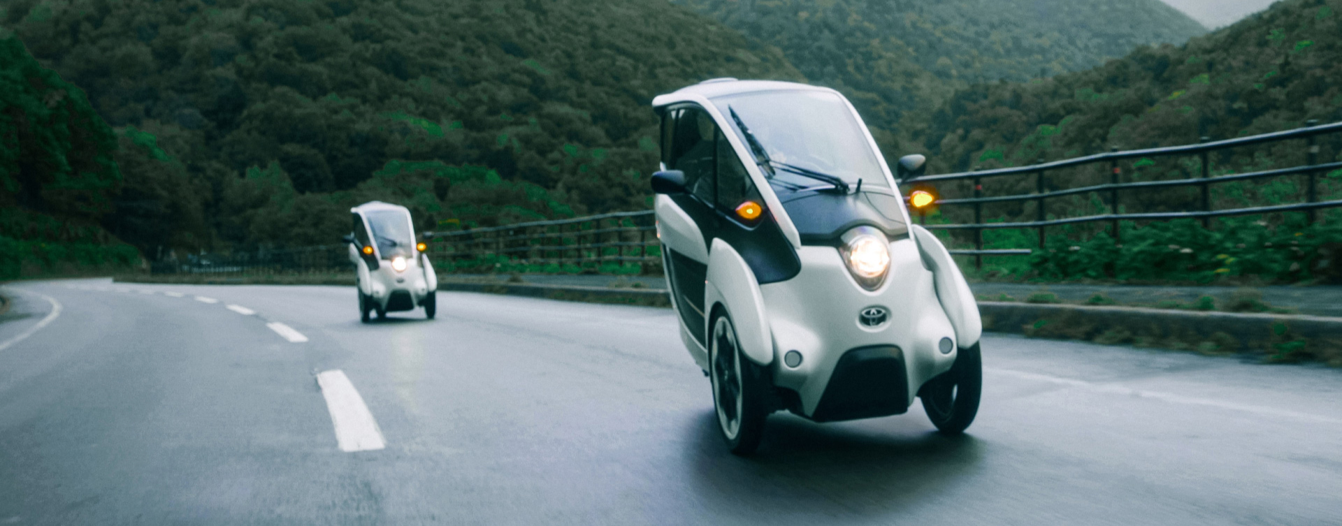 Two futuristic looking Toyota concept vehicles driving along a wet highway surrounded by lush green mountains.