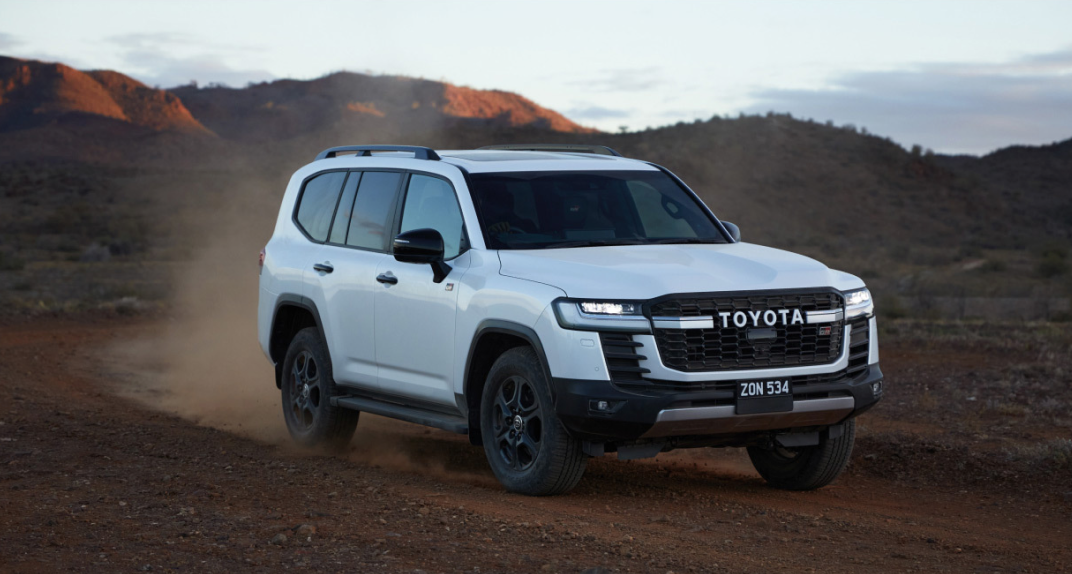 A white Toyota LandCruiser 300 traverses a dirt road in the orange Australian outback at dusk.