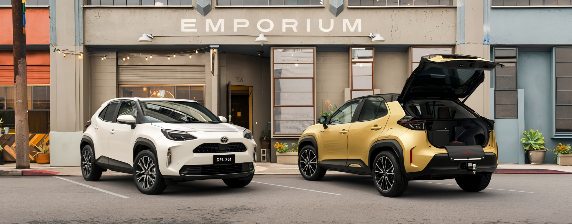  Two Yaris Cross parked in front of a grey urban building that says ‘EMPORIUM’. The left vehicle is white and facing away from the building, the right vehicle is gold and is facing towards the building with the boot open.