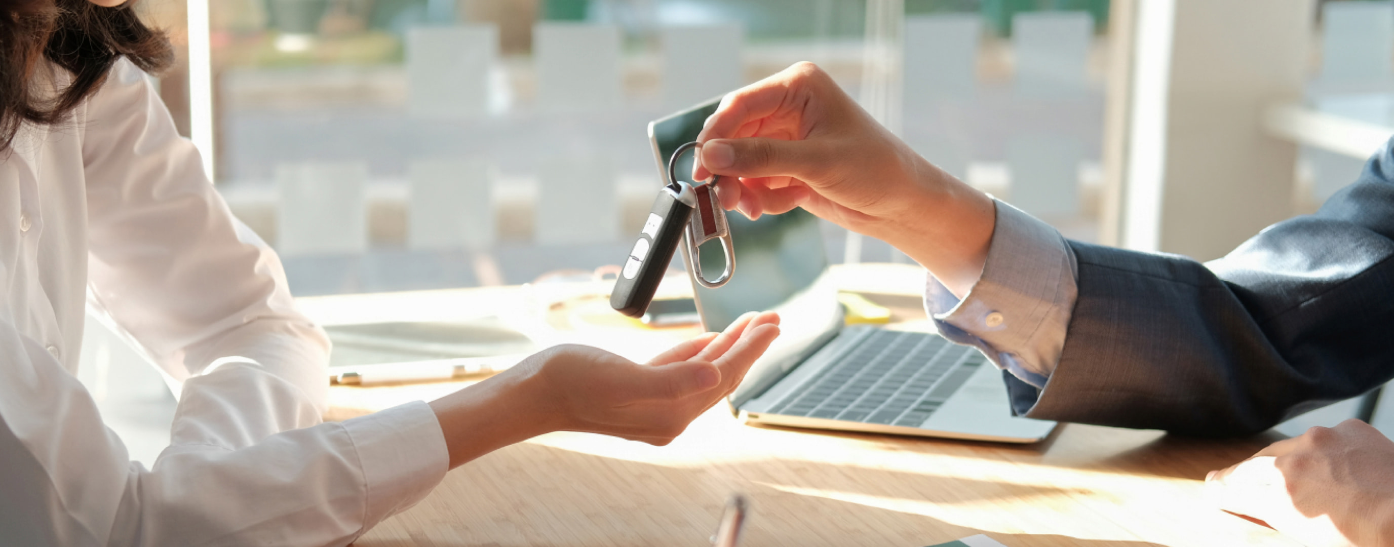 Close-up of one person handing car keys to another across a desk, with a laptop in the background.