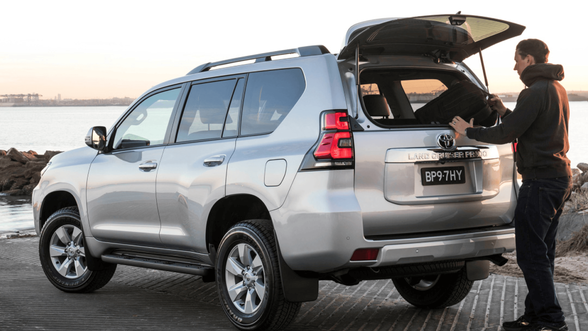 A man in a hoodie removing a bag from the open rear window of his Toyota Land Cruiser Prado, parked next to a bay with shipyards in the far distance.