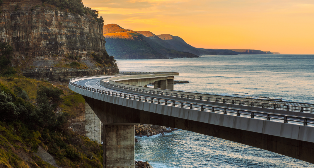 A curved coastal ocean highway, jutting out above the water on pylons, against a grassy rockface overlooking a bay.