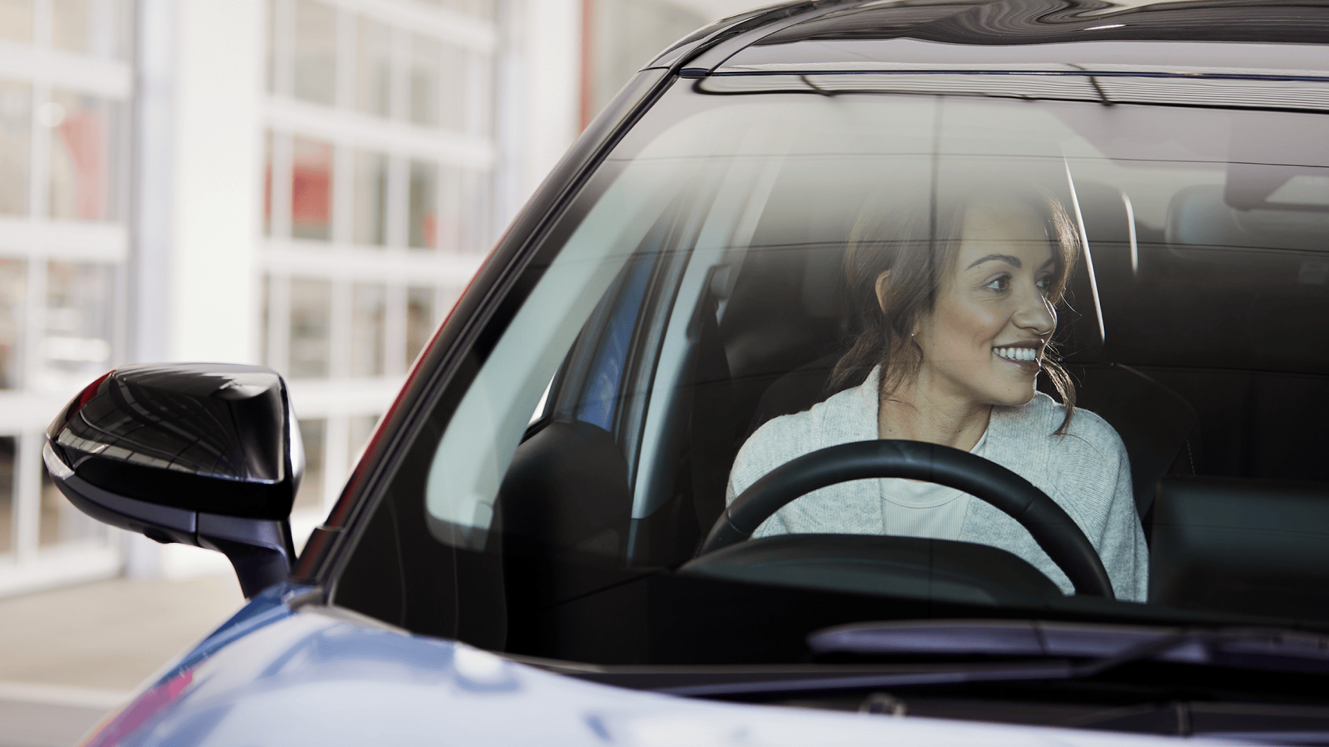 A smiling woman sits in the driver's seat of a Toyota, with the exterior of a service center visible through the windows.
