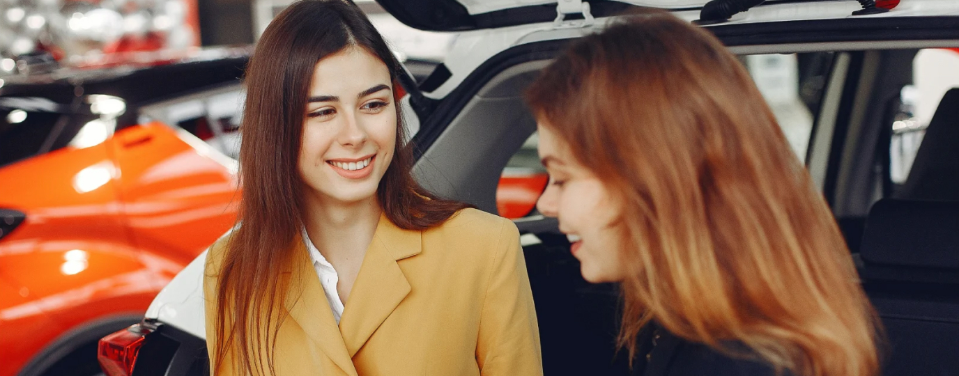 At a Toyota dealership, a Dealer smiles as she hands a Toyota customer a clipboard, the customer looking down at the clipboard’s contents happily.