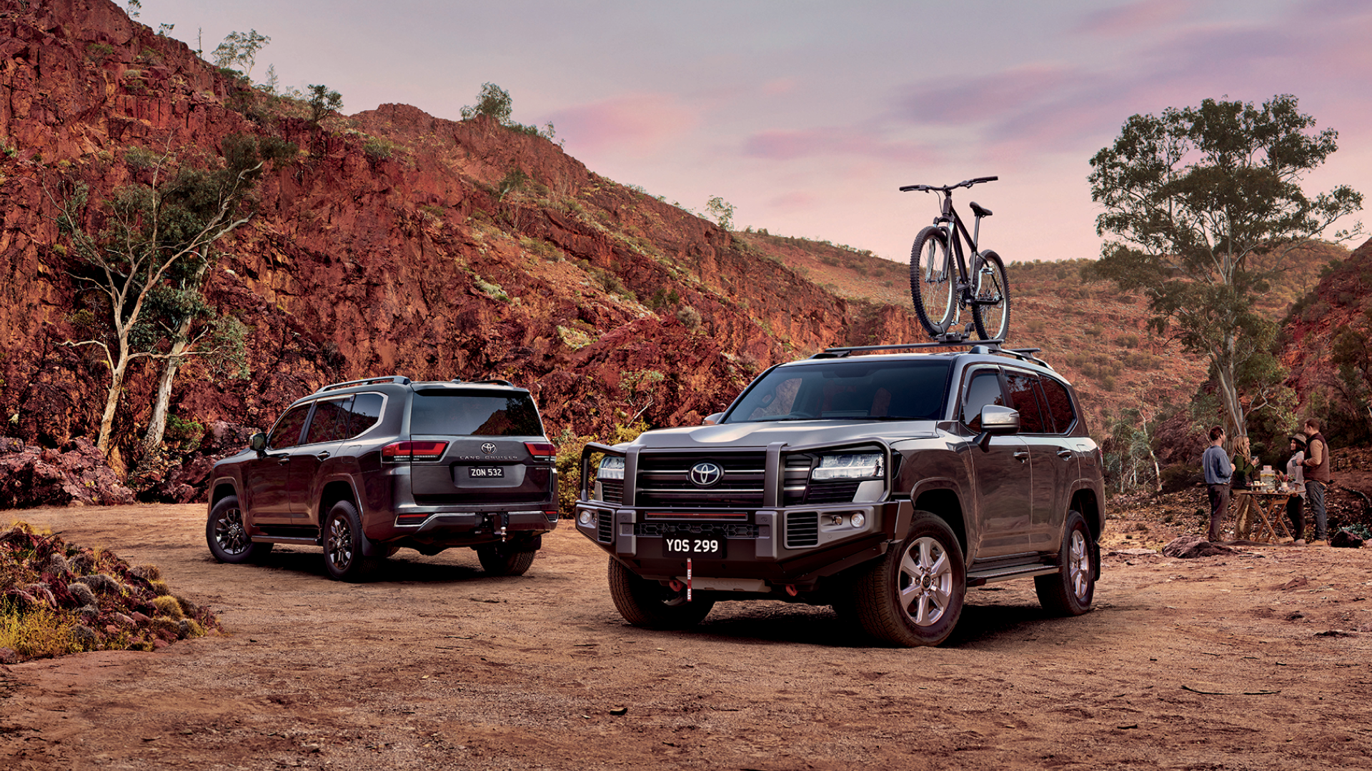 Two dark grey Toyota LandCruiser 300, parked in amongst the orange hills of the Australian outback, a group of campers socialising behind them. One LandCrusier is fitted with a Toyota Genuine Roof Rack, Bull Bar, LED Light Bar and Winch.