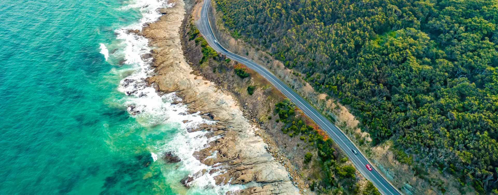 An aerial view of the Great Ocean Road, hugged by a rocky shoreline and a dense, green forest.