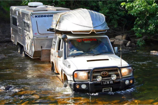 A 2007 landcruiser 70 Wagon GXL with a dinghy attached to the roof rack, driving through a shallow freshwater river in the outback while towing a caravan.