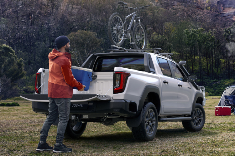 A man in an orange coat and beanie at a mountainous campsite, retrieving a blue esky out of the tray of a white HiLux
