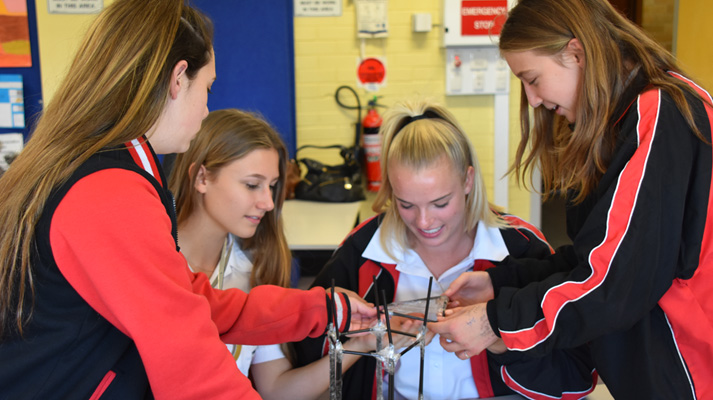 Four teenage girls focused intently, performing a group challenge involving building a tower using just black straws and sticky tape.