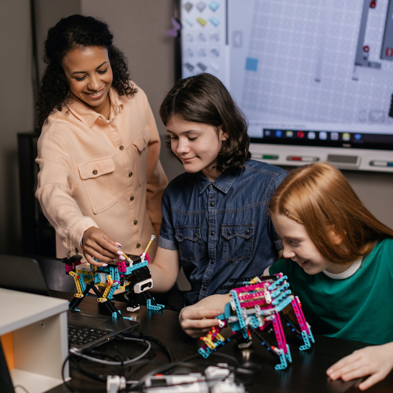 An ‘In2science’ mentor working together with two teenage girls to construct robots with LEGO Education SPIKE sets.