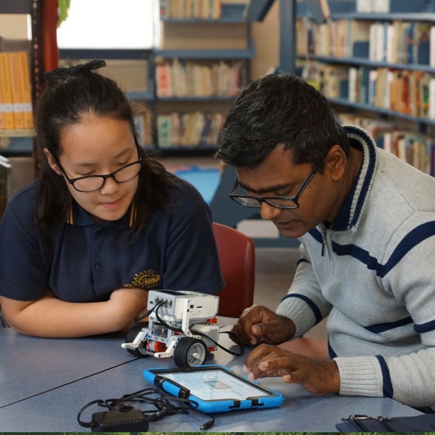 A teenage schoolgirl sits beside her teacher while he examines a small machine that she has constructed.