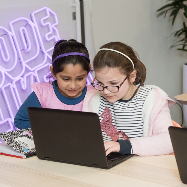 Two young girls participate in ‘Code Like a Girl’. The wall behind them is decorated with a purple neon sign, intently looking at what’s displayed on a laptop in front of them.
