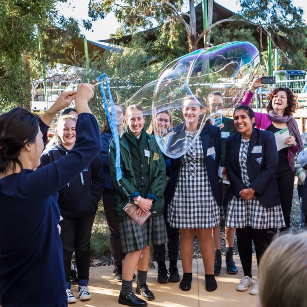 A group of school girls outside on a sunny afternoon in the shade, enjoying a woman demonstrating how to make giant soap bubbles out of bubble mix.