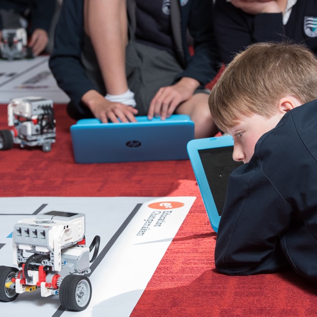 Young school children sat on a red carpet, each with a blue HP laptop and a small robotic electronic.