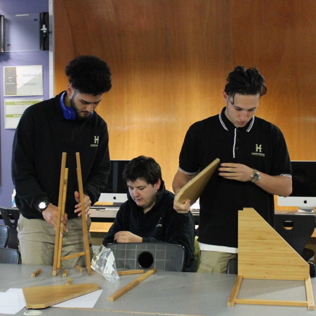 A group of three teenage schoolboys in a classroom full of Mac computers, working together to construct something out of pre-supplied cut pieces of wood. 