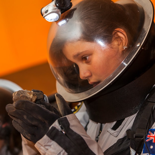 A young girl wears an astronaut-like suit and examines a mineral-like rock in her gloved hands.