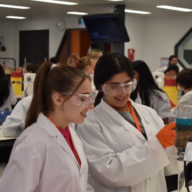 A busy school science laboratory, with two youthful girls wearing lab coats and safety specs, examining a plastic bottle full of layers of different minerals, and a blue liquid.