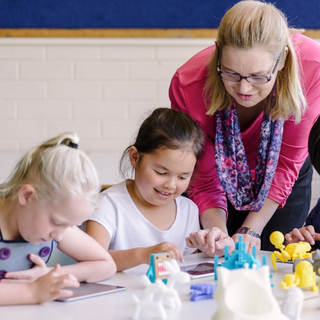 Two preschool-aged girls with a preschool teacher overlooking their learning activity. The girl in the middle of the other preschooler and the teacher are smiling as she completes her task.