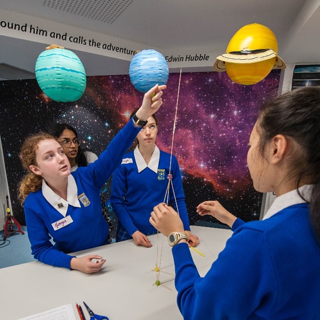 Three female high-school students wearing vivid royal blue uniforms participate in a tower-building team challenge using only pieces of uncooked pasta and putty. Their female teacher is observing from behind.