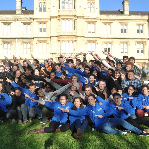 A large group of youths making the same choreographed pointing gesture into the sky, during the day out the front of a large mansion-like building.