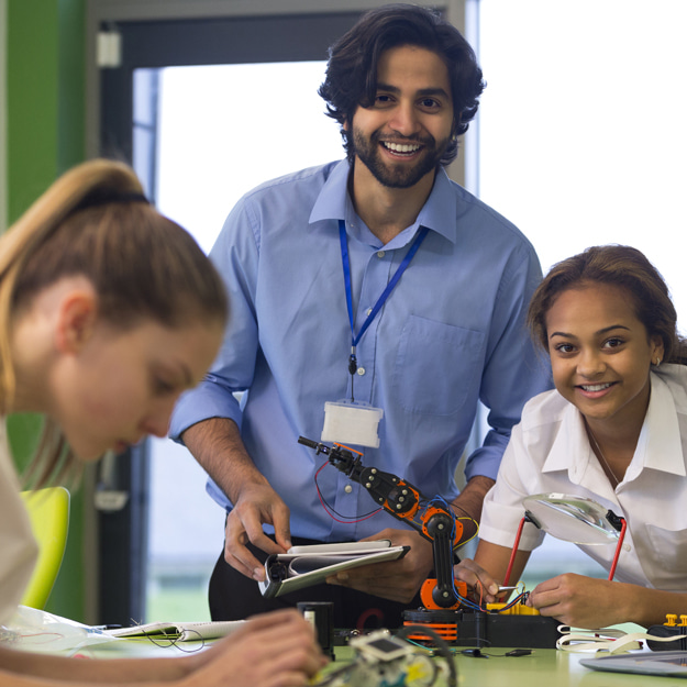 Three youthful people working on miniature robotics, one in the forefront out of focus, two in the background in clear focus, a male holding a notebook and smiling ahead, and the female smiling ahead whilst connecting wires attached to a miniature robotic arm.