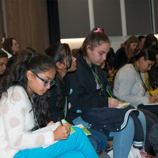 Young women taking notes during a presentation in a dimly-lit auditorium.