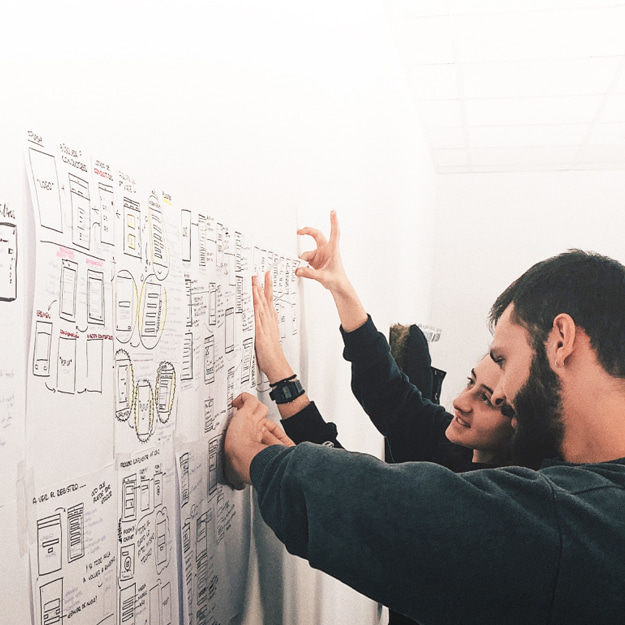 A male and a female address heavily annotated handwritten posters stuck up on a white wall in deep discussion about the content.