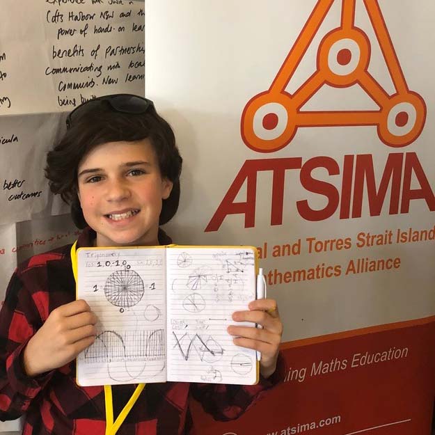 A young boy with luscious brown hair and sunglasses on top of his head, proudly holding up a notebook full of hand-drawn scientific diagrams. The boy stands in front of an ASTIMA (Aboriginal and Torres Strait Islander Mathematics Alliance) banner. 