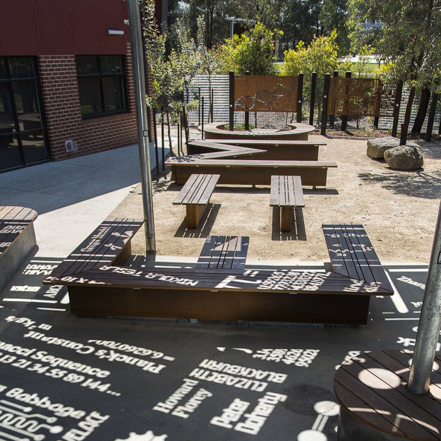 Wooden benches in a shaded recreational area at a primary school, stencils cut into the roof of the cover to project sunlight into the benches in the shapes of letters and words.
